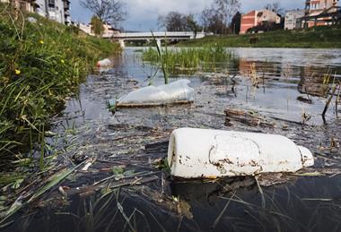 Close-up_of_plastic_bottles_polluting_the_river_with_a_bridge_and_buildings_in_a_blurry_background_(51142599174)